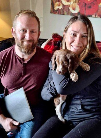 Couple smiles, holding a puppy with brown and white fur indoors, painting on the wall.
