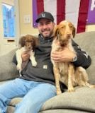 Man smiling on a couch holding two dogs; one small and fluffy, the other large with long ears; US flag in the background.