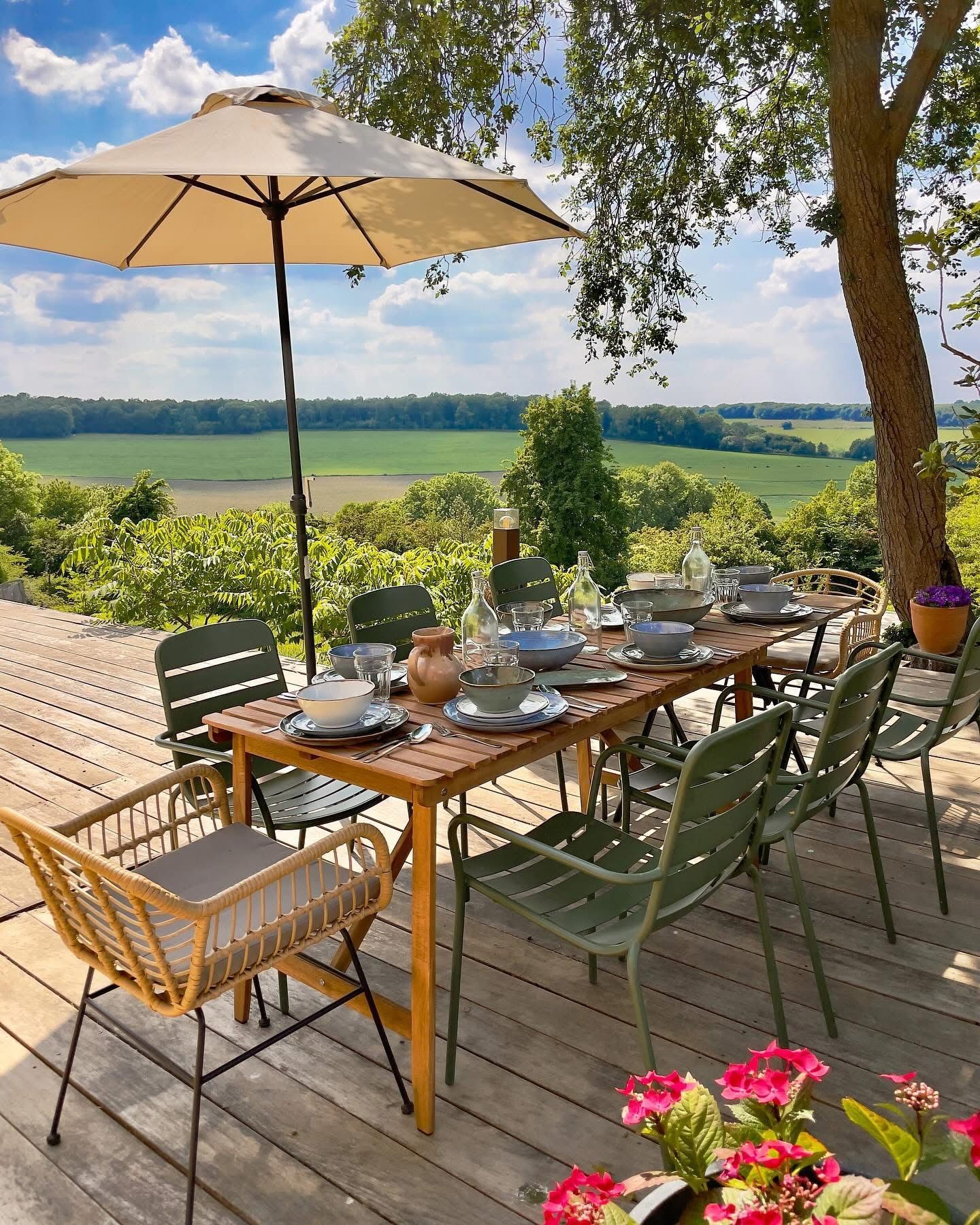 Outdoor dining table set for a meal on a wooden deck with a scenic view, under a beige umbrella.