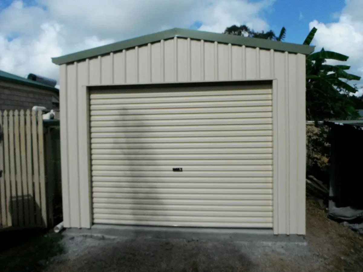 Tan garage with a roller door, behind a wooden fence, under a cloudy sky -Shed Constructions (QLD) In Gin Gin, QLD