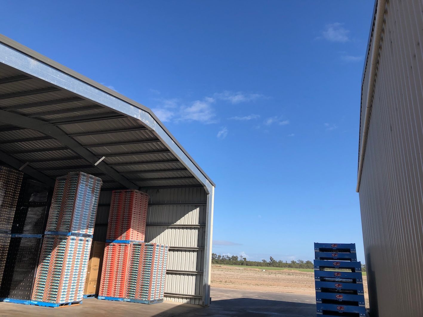 Warehouse exterior with pallets of packaged goods under a metal roof, blue sky above.