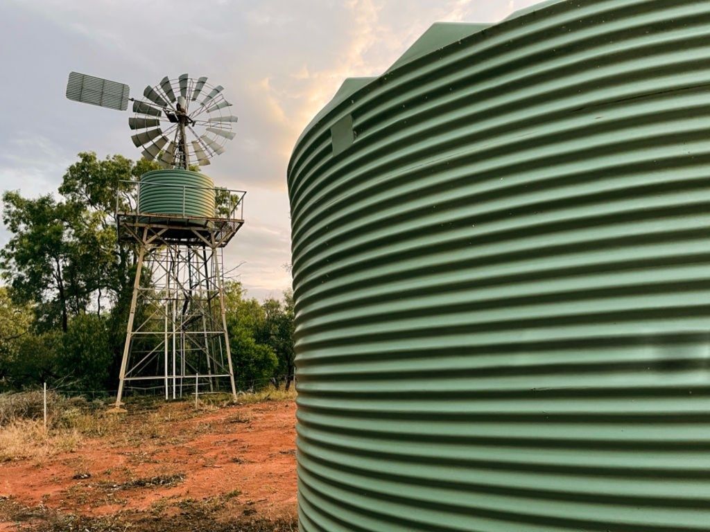 Green corrugated water tank beside a windmill with a water tank on a tower in a dry, grassy landscape — Shed Constructions (QLD) In Gin Gin, QLD