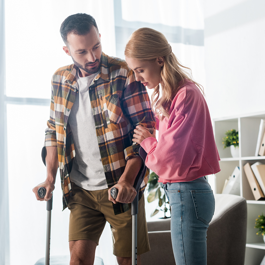 A woman is helping a man with crutches walk in a living room.