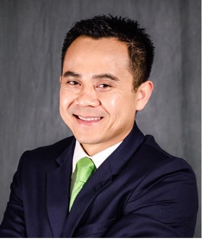 Smiling man in navy suit and green tie, against a gray background.