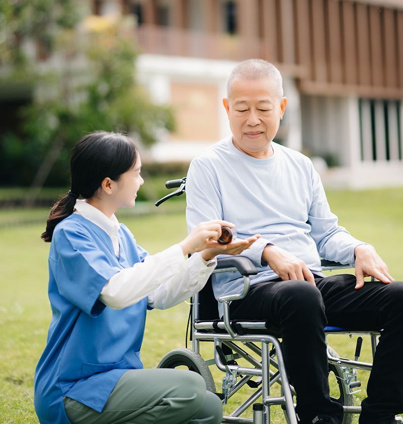 Caregiver gives medicine to an elderly man in a wheelchair outdoors on grass.