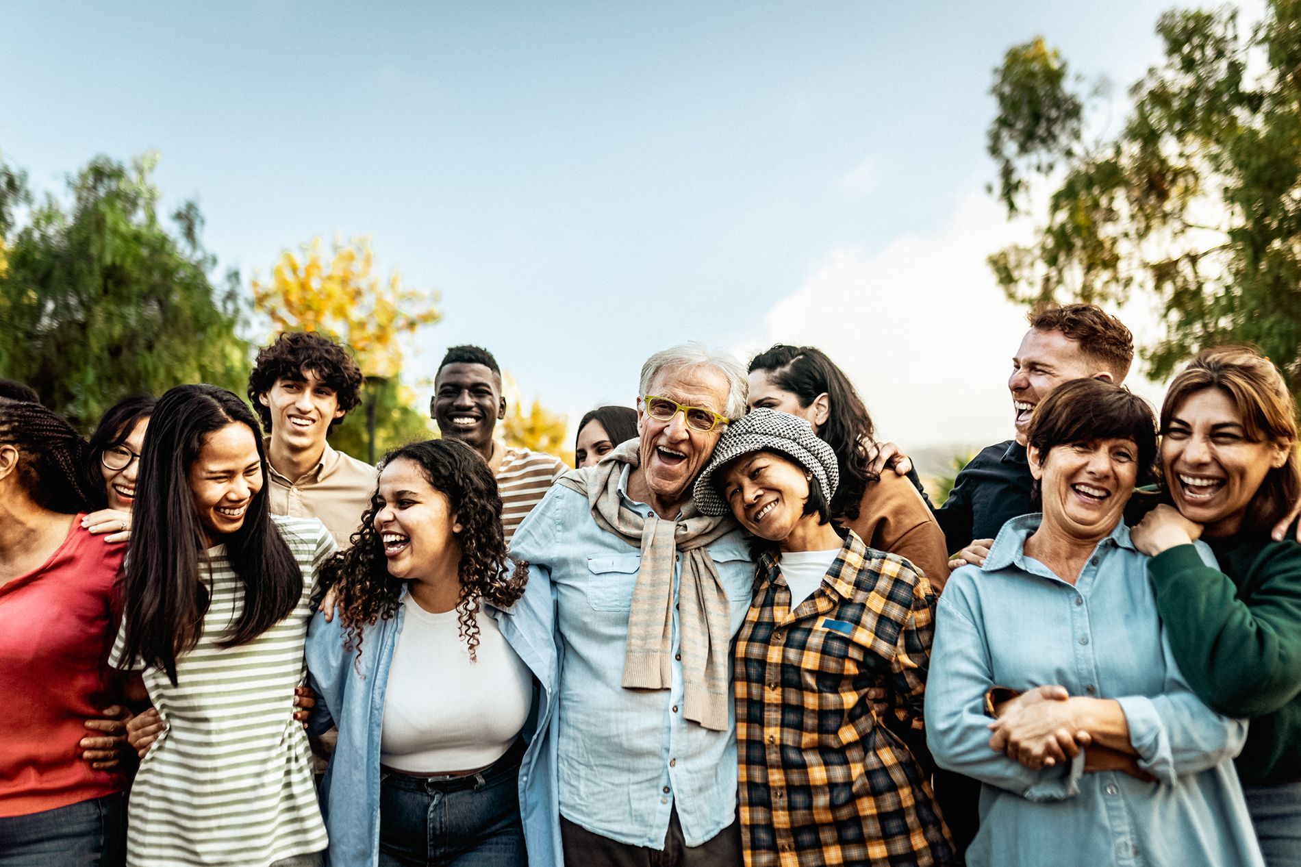 Group of diverse people smiling and embracing outdoors, with trees and a blue sky.