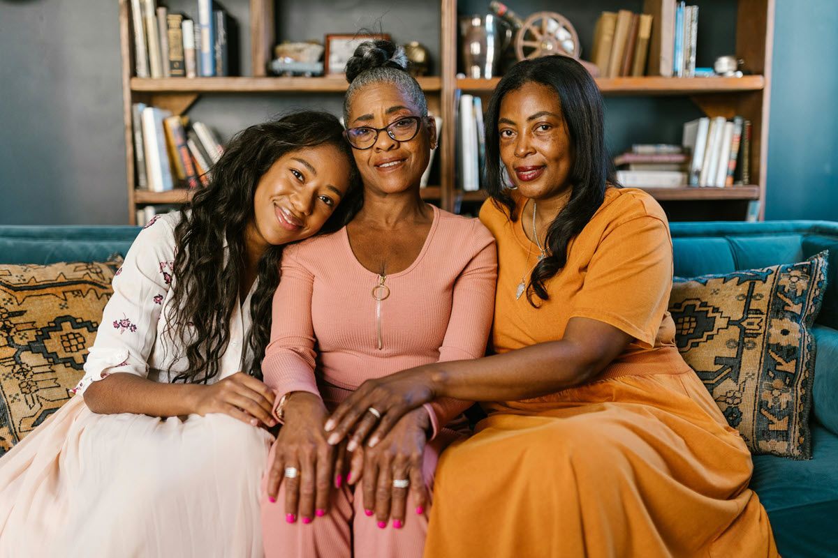 Three Black women smiling on a blue couch in a room with a bookshelf. One rests her head on another's shoulder.