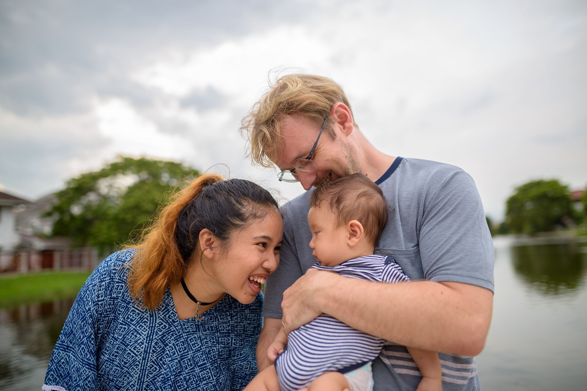 Family of three: woman, man holding baby near water, smiling.