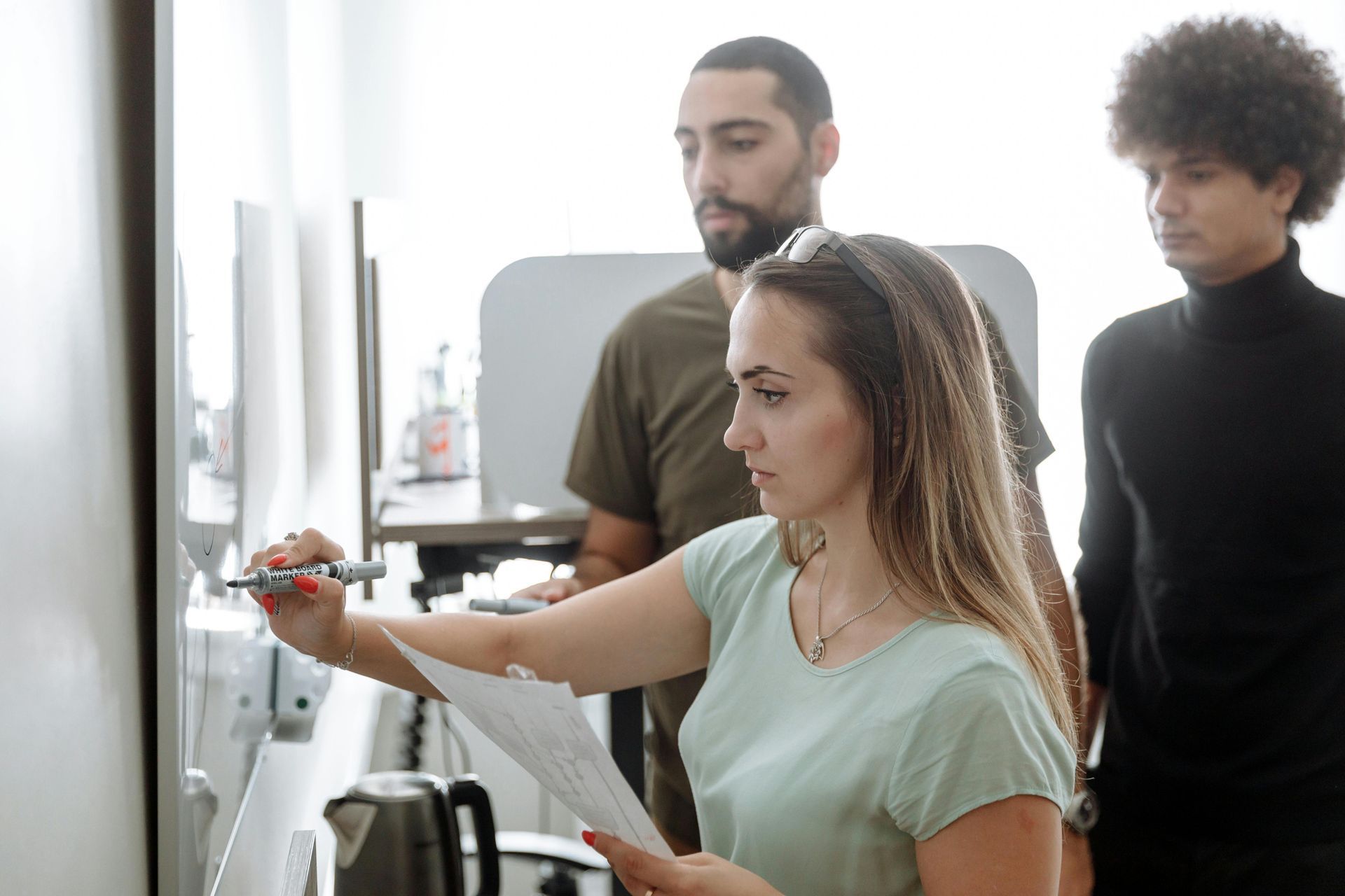 Three people, two men and one woman, at a whiteboard, the woman is writing.