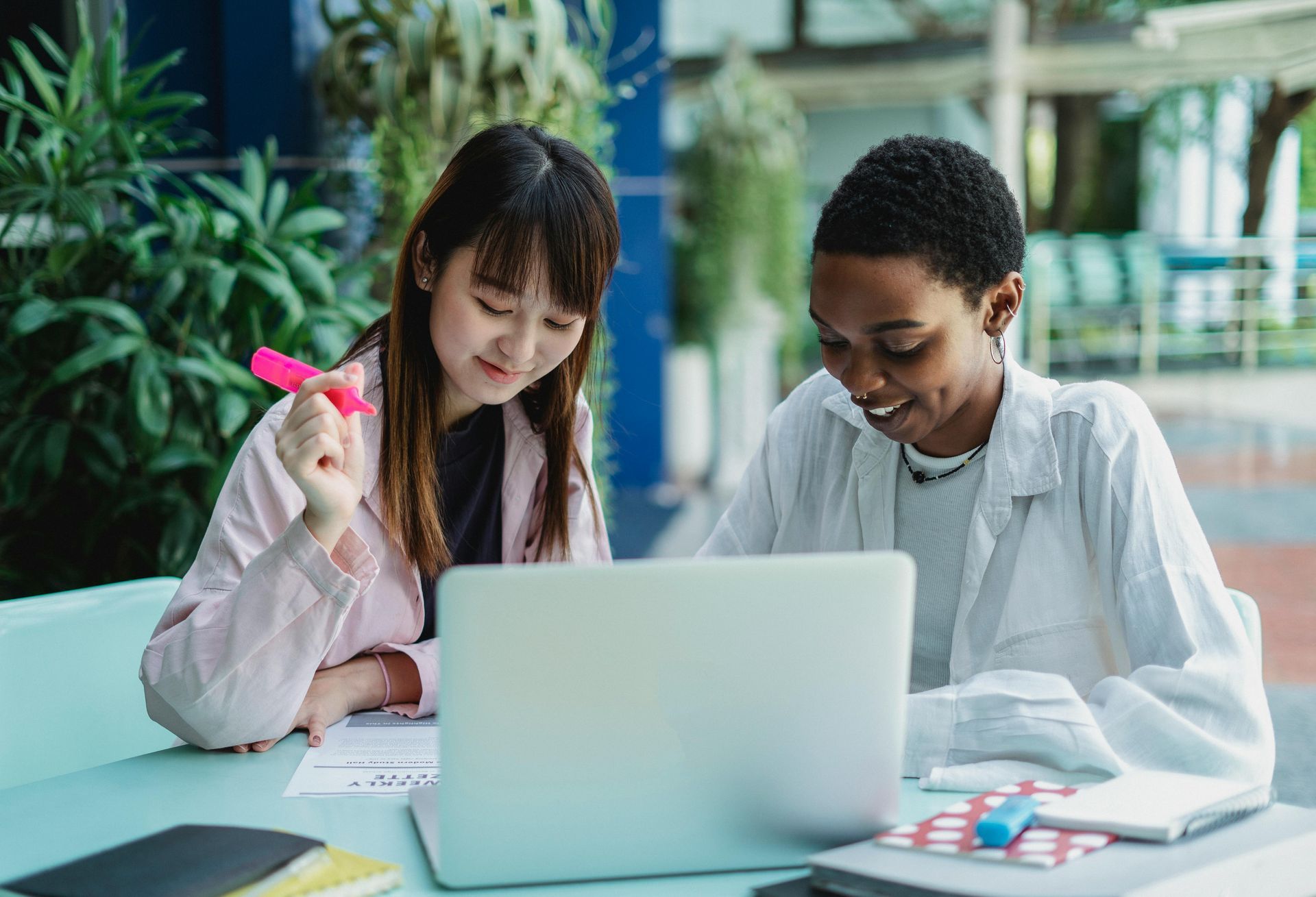 Two women at a table looking at a laptop, one highlights notes. Outdoor setting, both smiling.