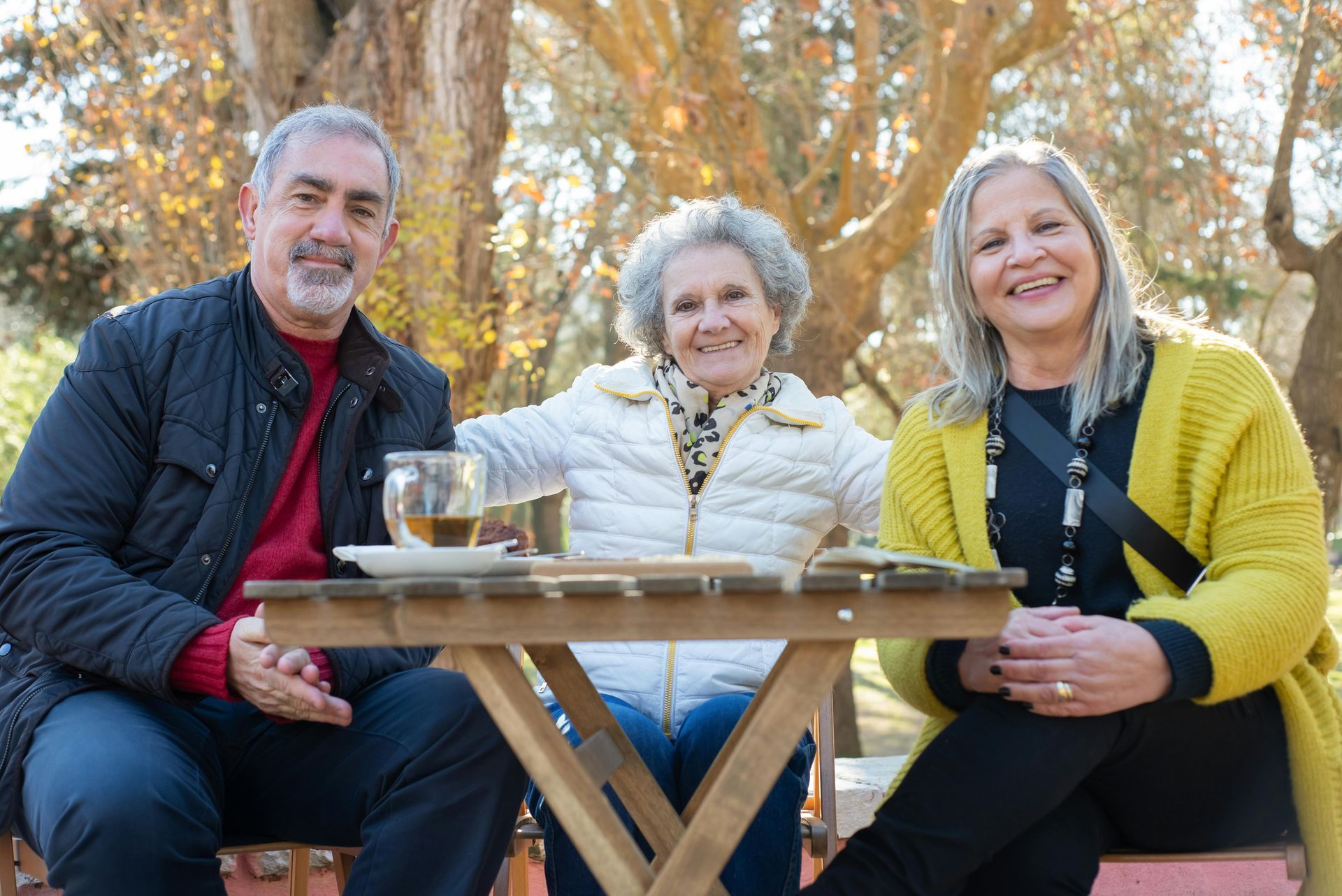 Three smiling seniors seated at a table outdoors. Man in a red sweater, two women in white and yellow sweaters.