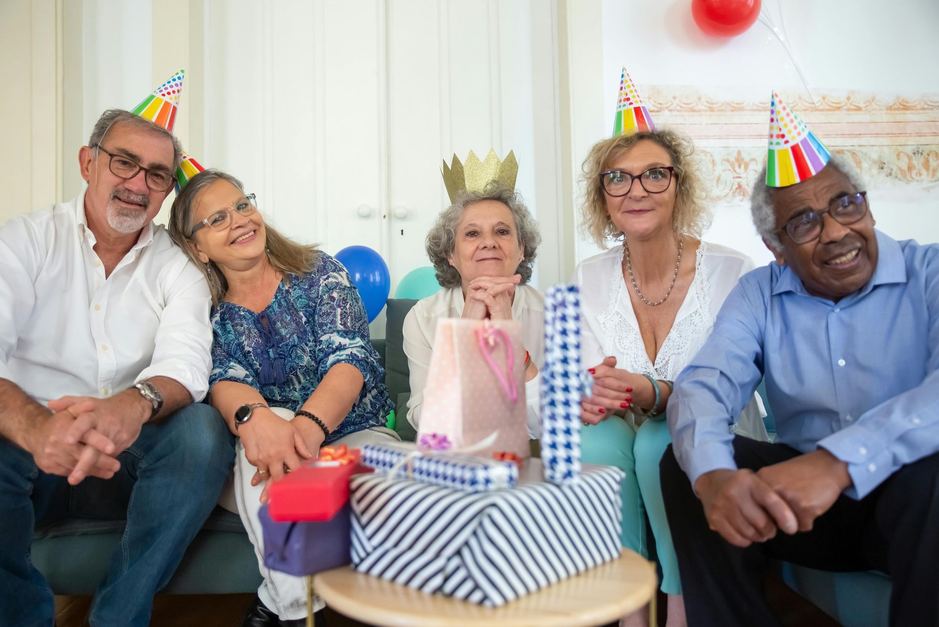Group of five smiling seniors celebrating a birthday, wearing party hats, surrounded by gifts.