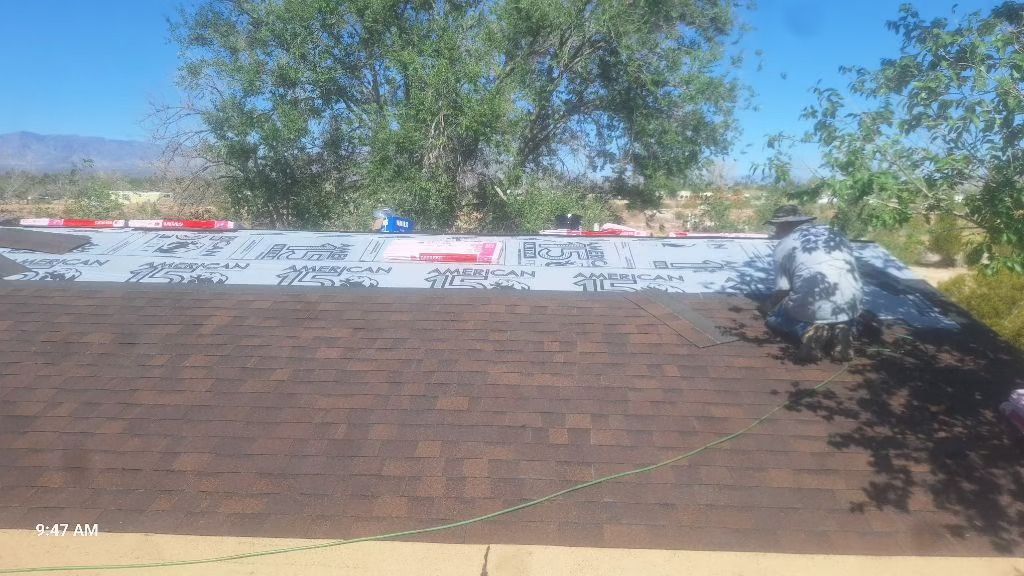 Roof partially covered in brown shingles, with exposed underlayment and clear blue sky.