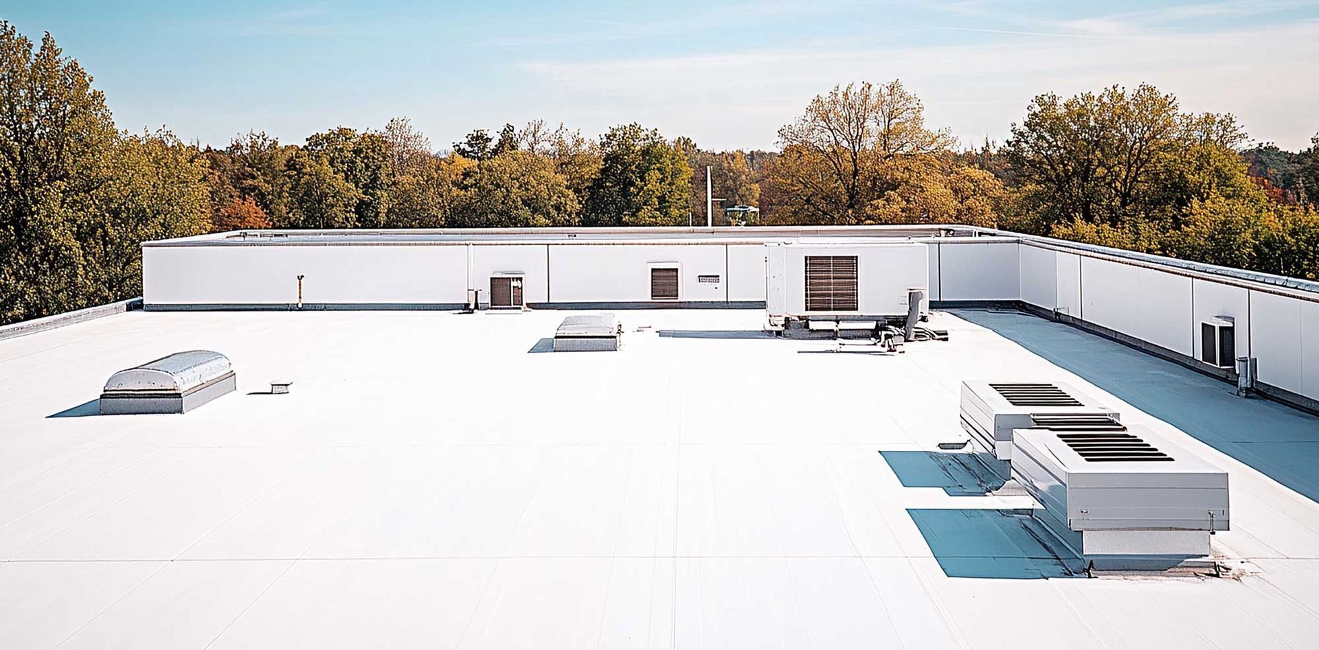 A white commercial flat roof with equipment, a building, and trees in the background under a blue sky.