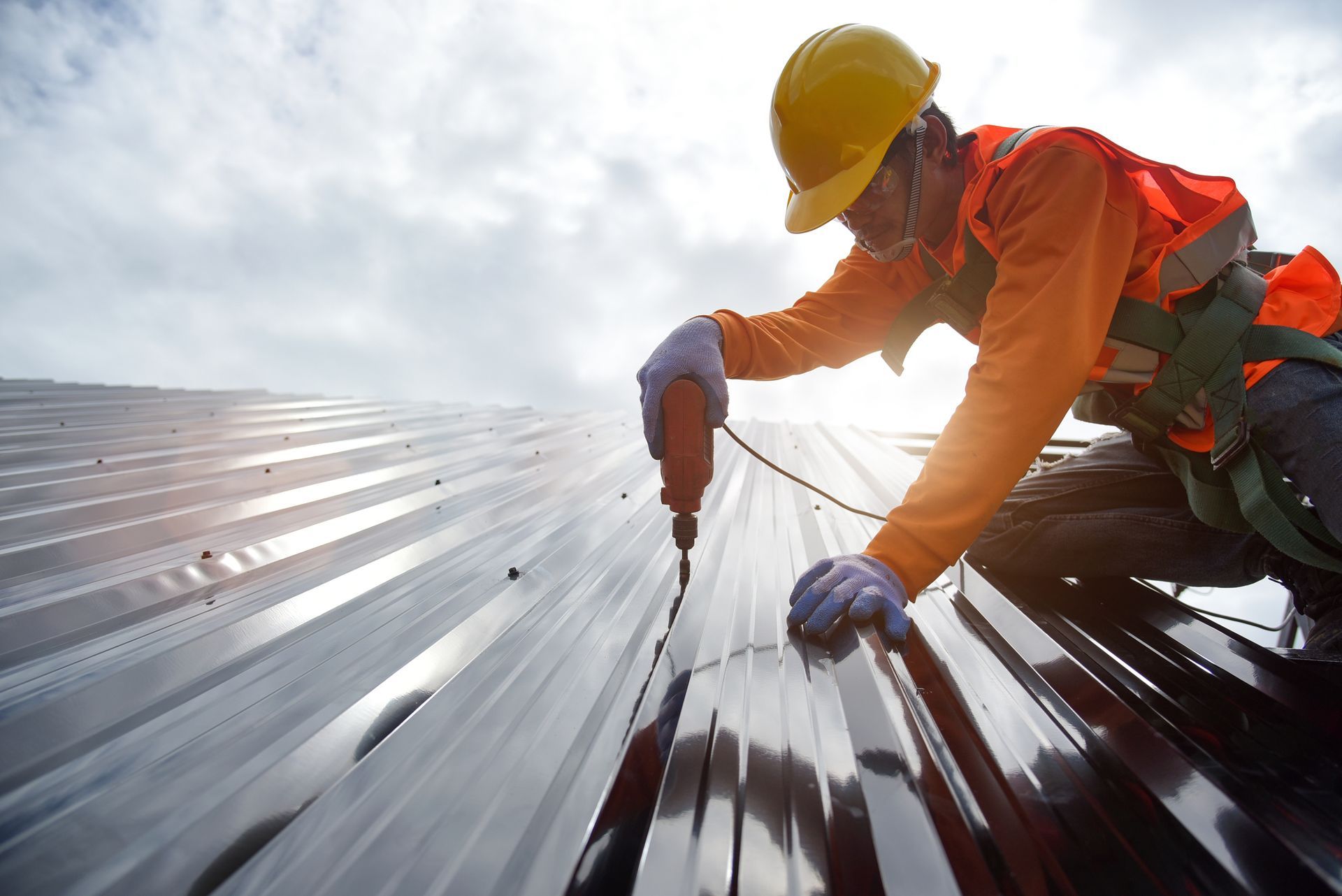 Roofer in safety gear using a power drill on a metal roof under a cloudy sky.