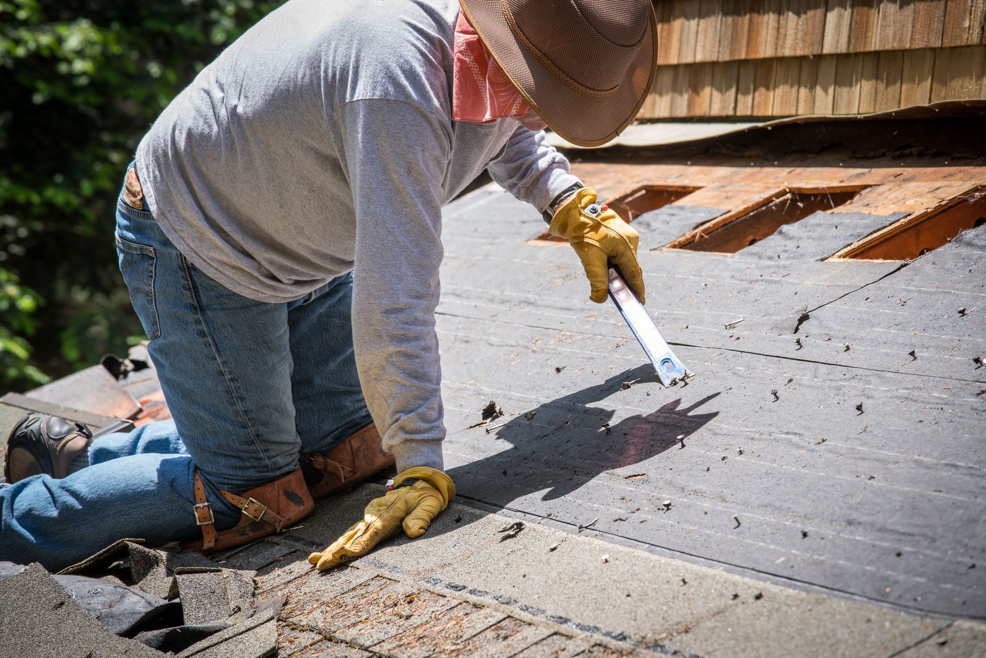 Man on a roof repairing shingles; wearing hat, gloves and jeans, using a scraper.