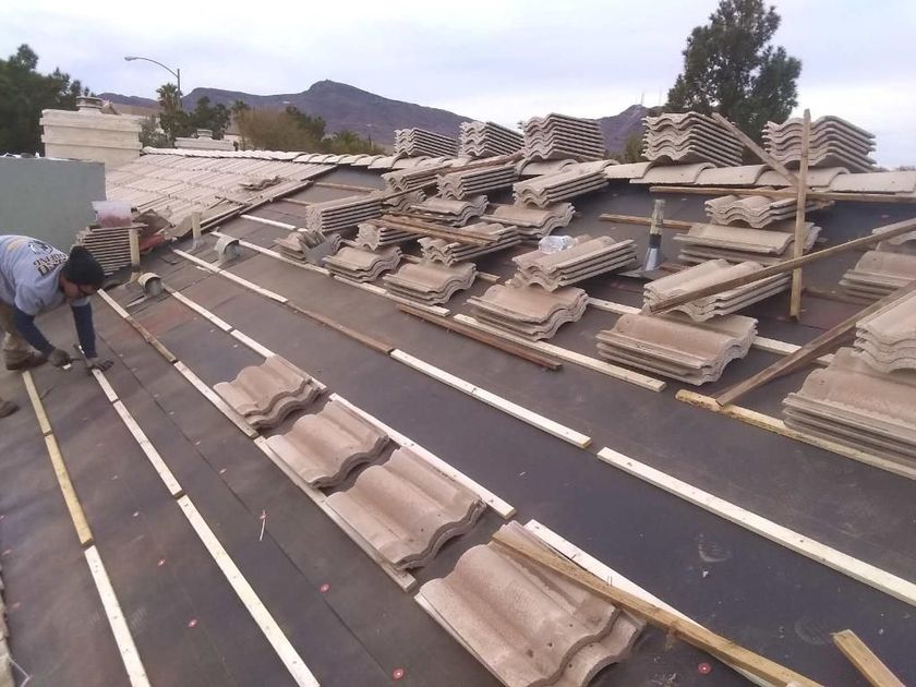 Roofer installing tiles on a dark roof with a mountain backdrop; stacks of tiles and lumber are present.