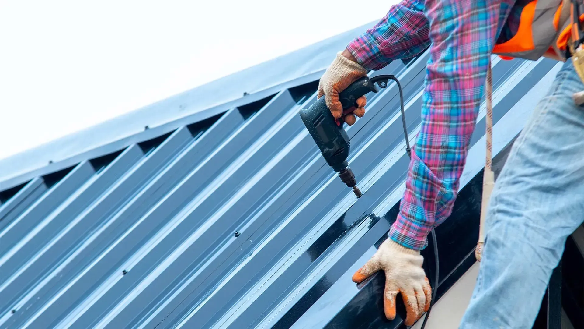 Roofer installing metal roofing with a drill; wearing gloves and safety harness.