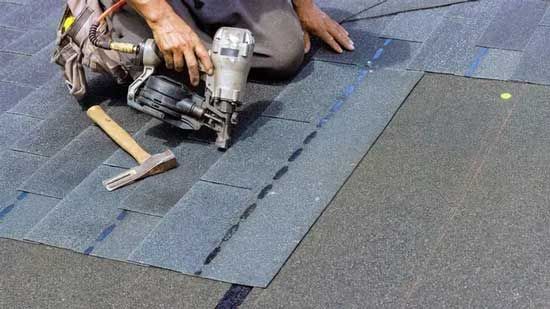 Roofer kneeling, using a nail gun to install dark asphalt shingles on a roof.