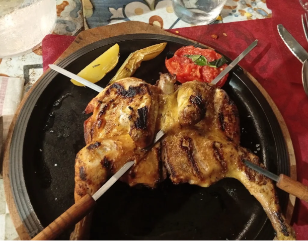 Pollo grigliato su un piatto nero con pomodoro, limone e patate, infilzato con i coltelli.