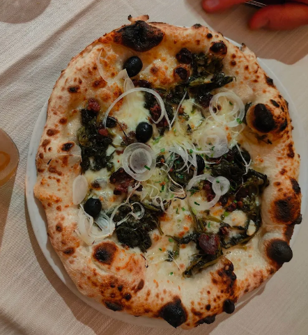 Pizza su un piatto bianco, guarnita con verdure verdi, cipolle e olive nere.