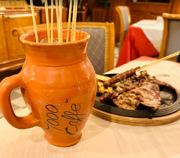 Una tazza di ceramica arancione con degli spiedini è appoggiata accanto a un piatto di carne al tavolo di un ristorante.