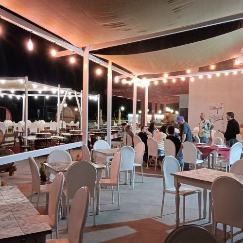 Ristorante all'aperto con tavoli e sedie bianchi, luci appese in alto, gente che cena.