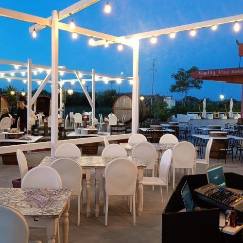Zona pranzo all'aperto con tavoli e sedie bianchi, sotto un pergolato bianco illuminato, al tramonto.
