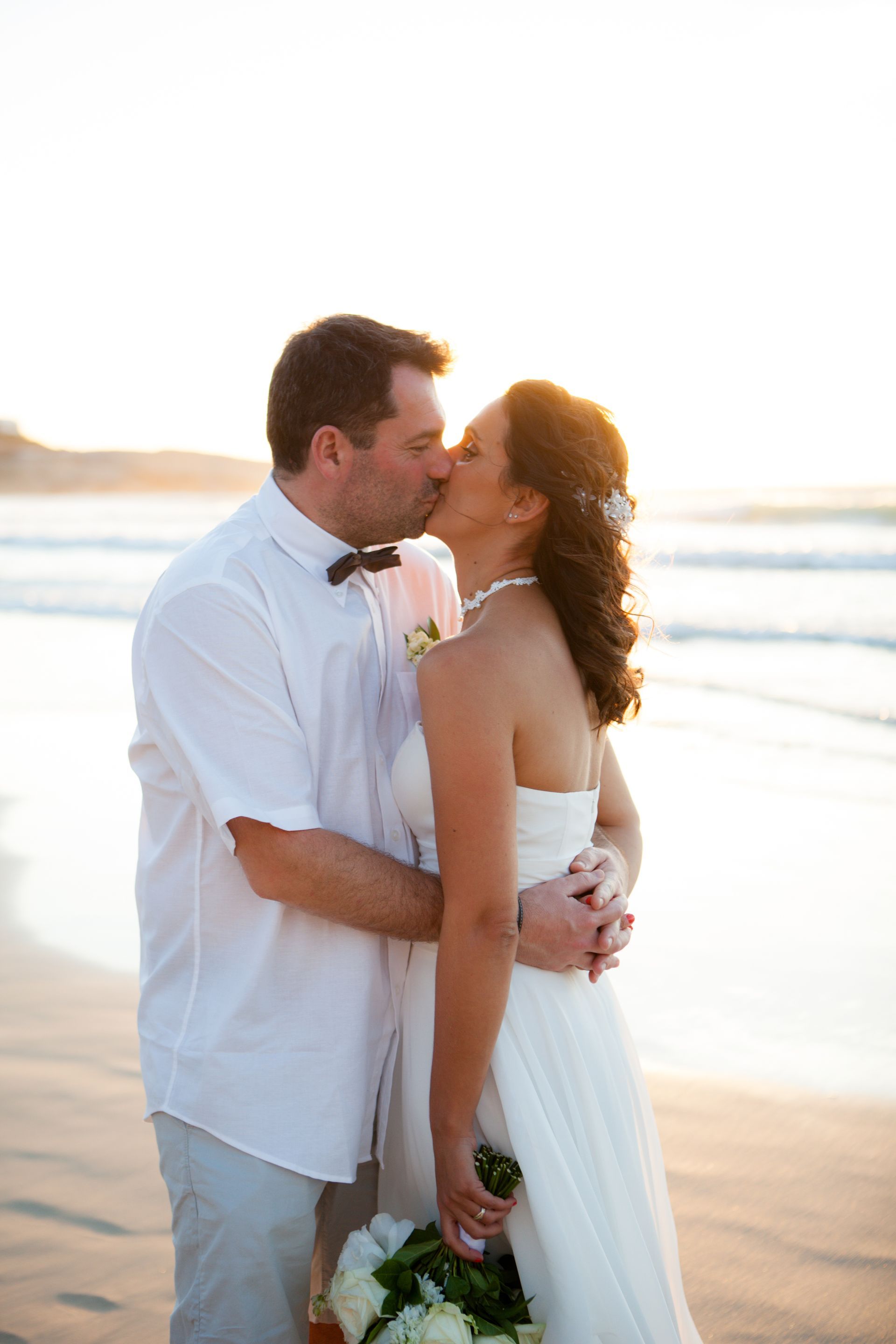 A bride and groom kissing on the beach at sunset.