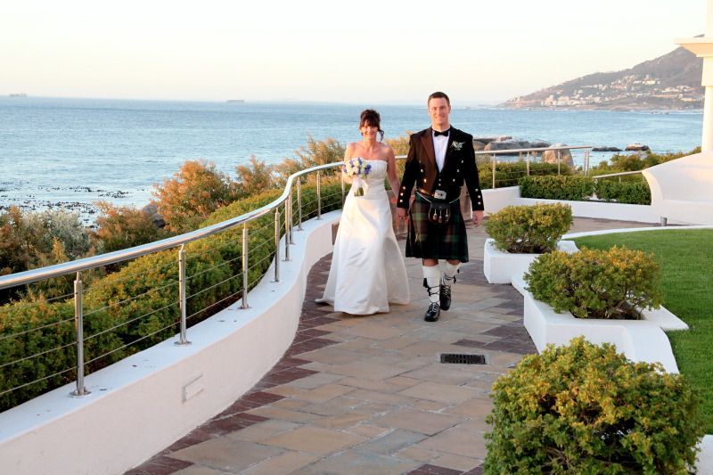 A bride and groom are walking down a path near the ocean