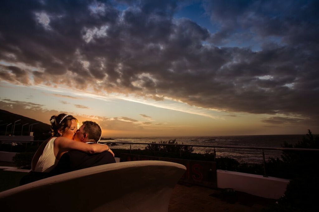 A bride and groom are sitting on a bench watching the sunset over the ocean.