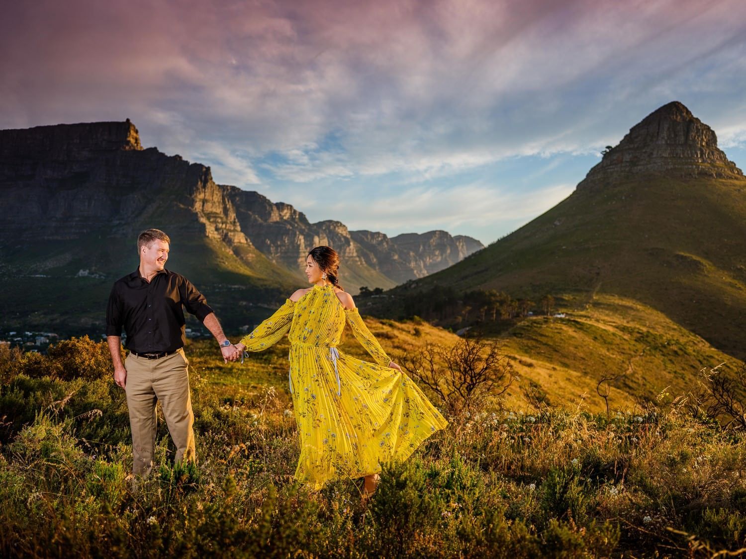 A man and a woman are holding hands in a field with mountains in the background.