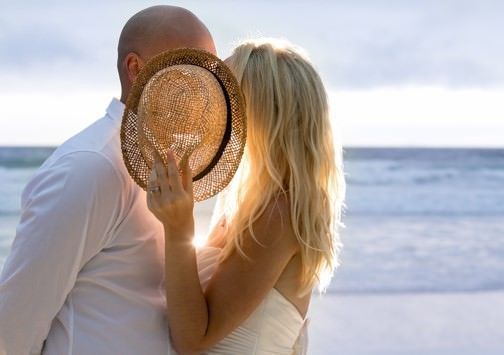A man and a woman are kissing on the beach while the woman covers her face with a hat.