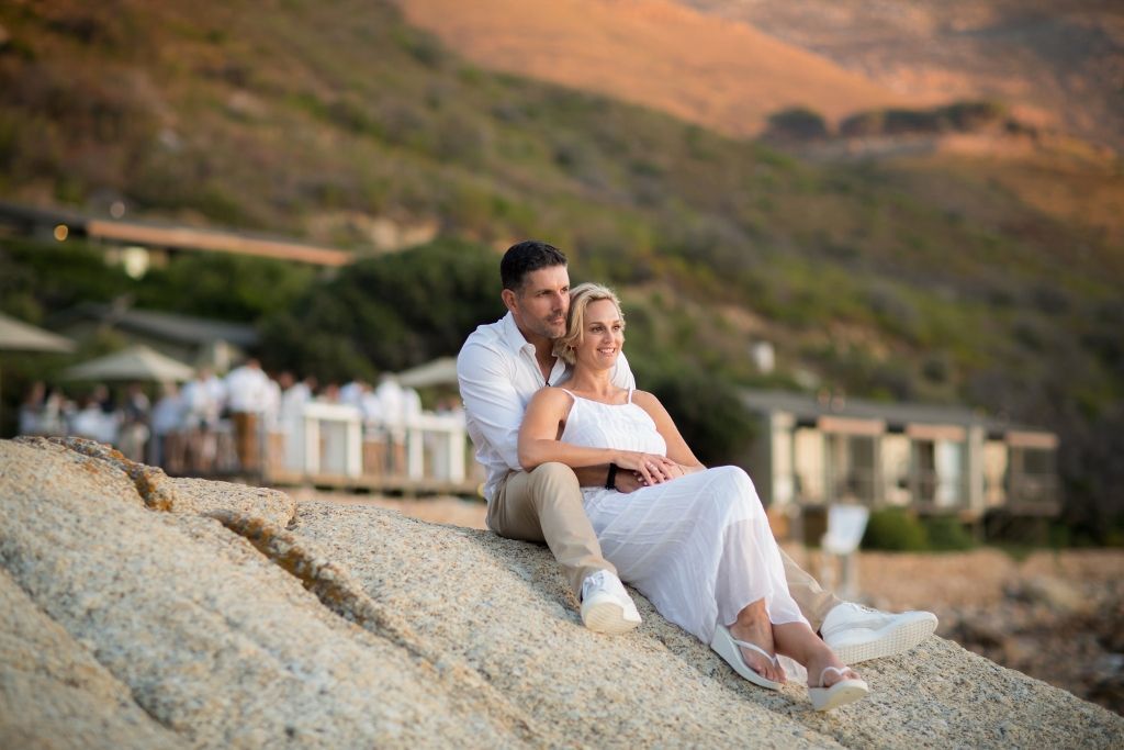 A bride and groom are sitting on a rock on the beach.