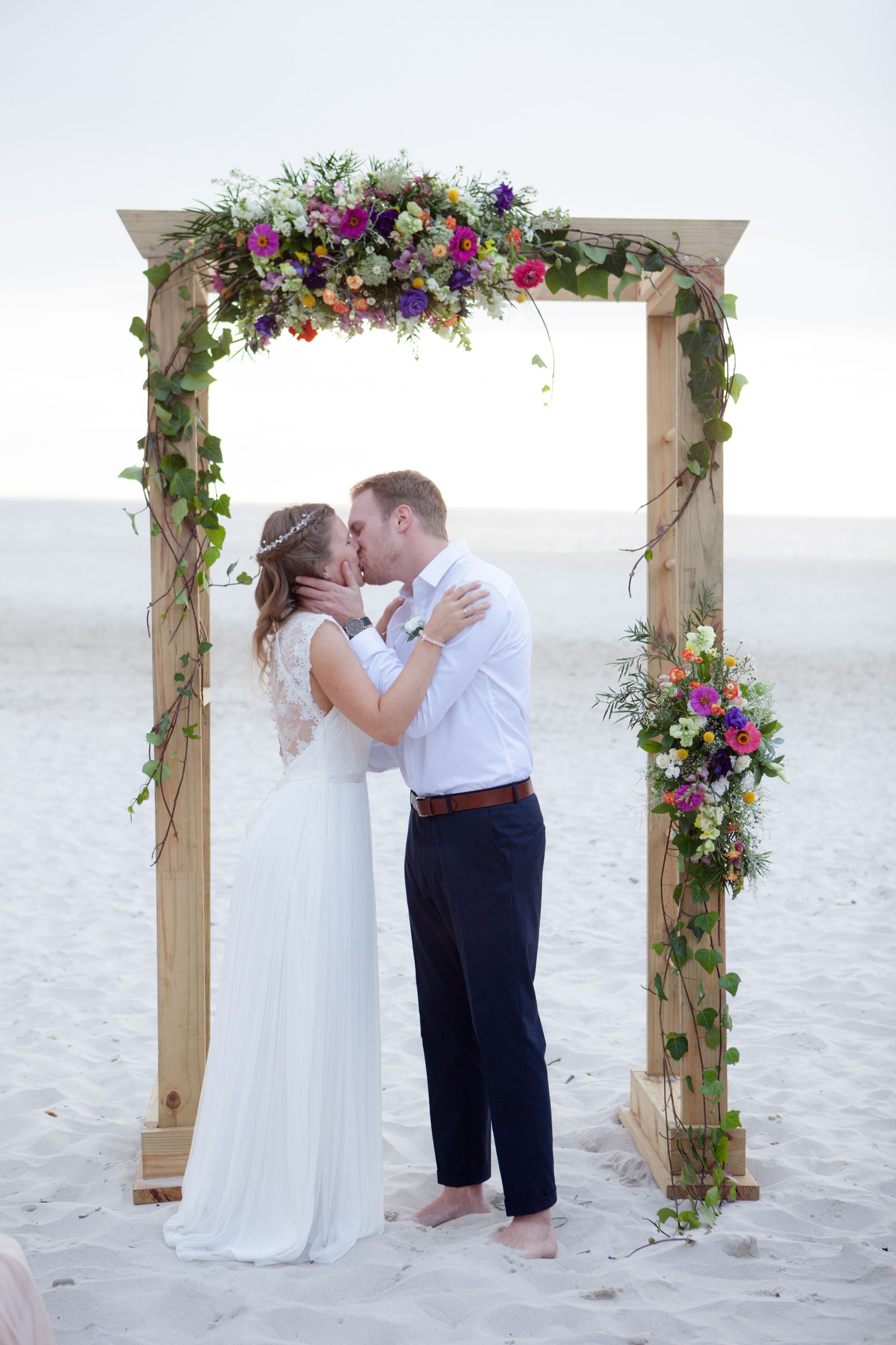 A bride and groom are kissing under a wooden arch on the beach.