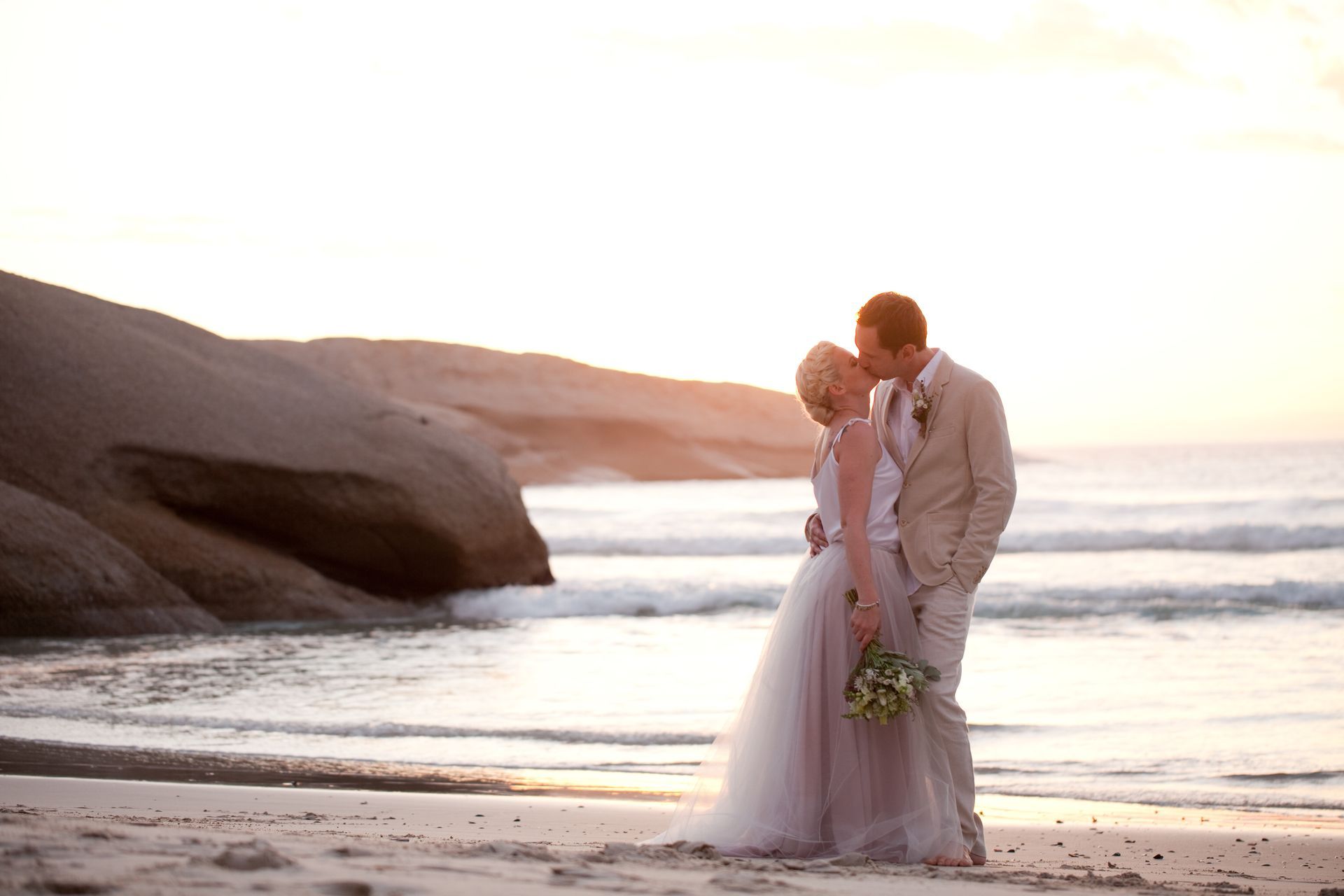 A bride and groom are kissing on the beach at sunset.