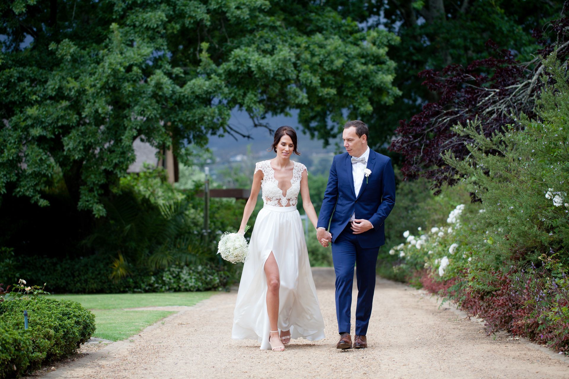 A bride and groom are walking down a dirt path holding hands.