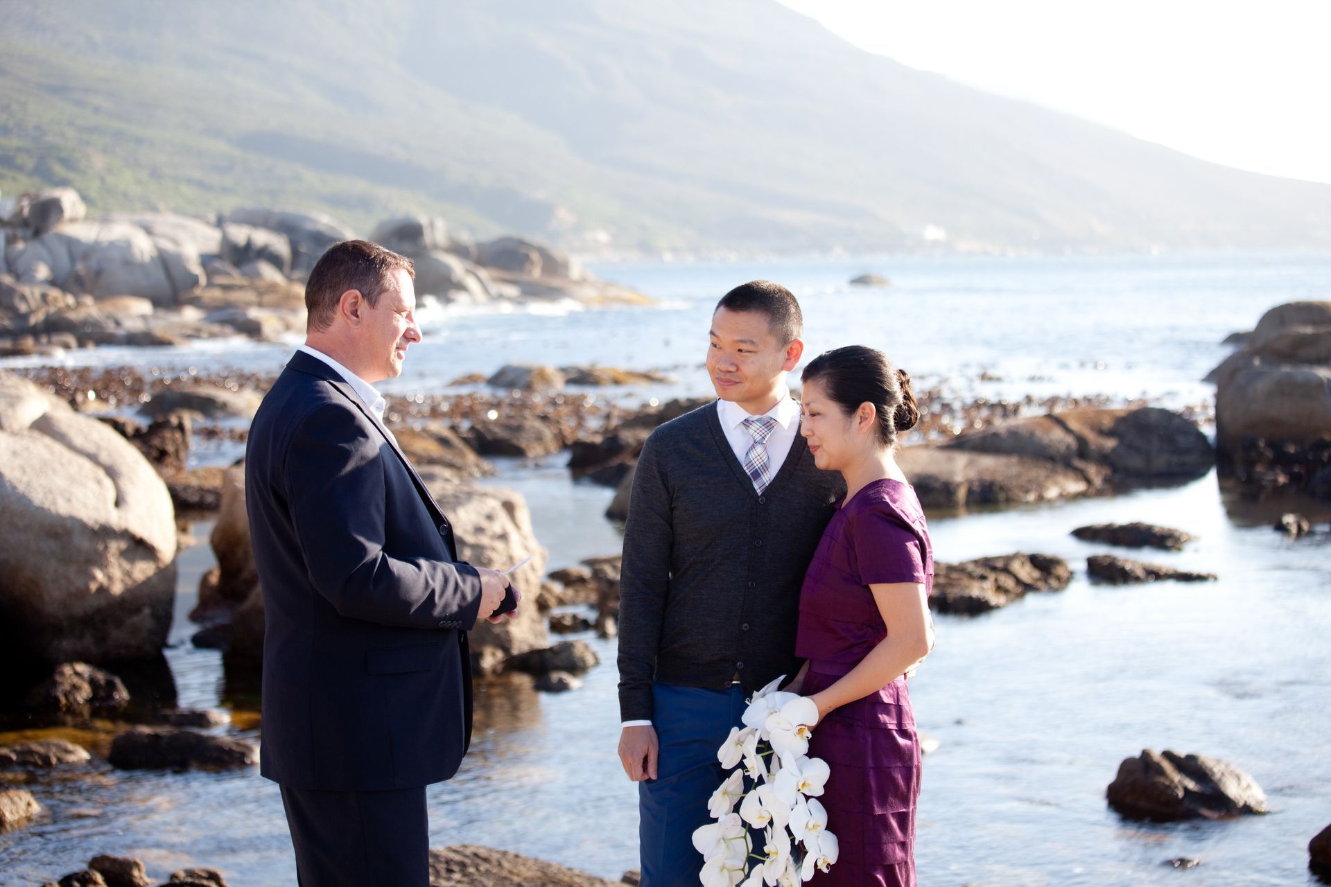 A man in a suit is giving a wedding ceremony to a bride and groom on the beach.
