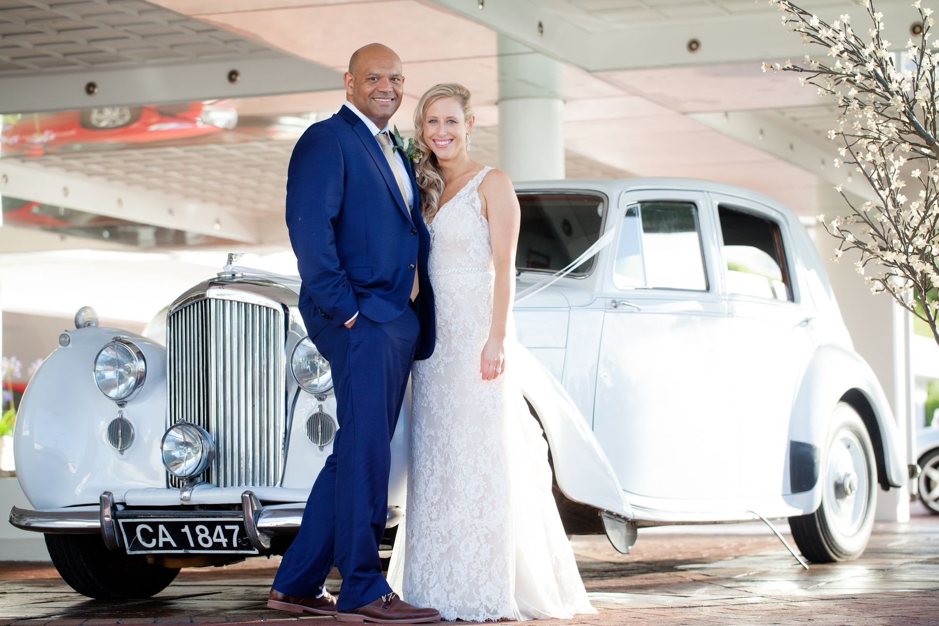 A bride and groom are posing for a picture in front of a white car.