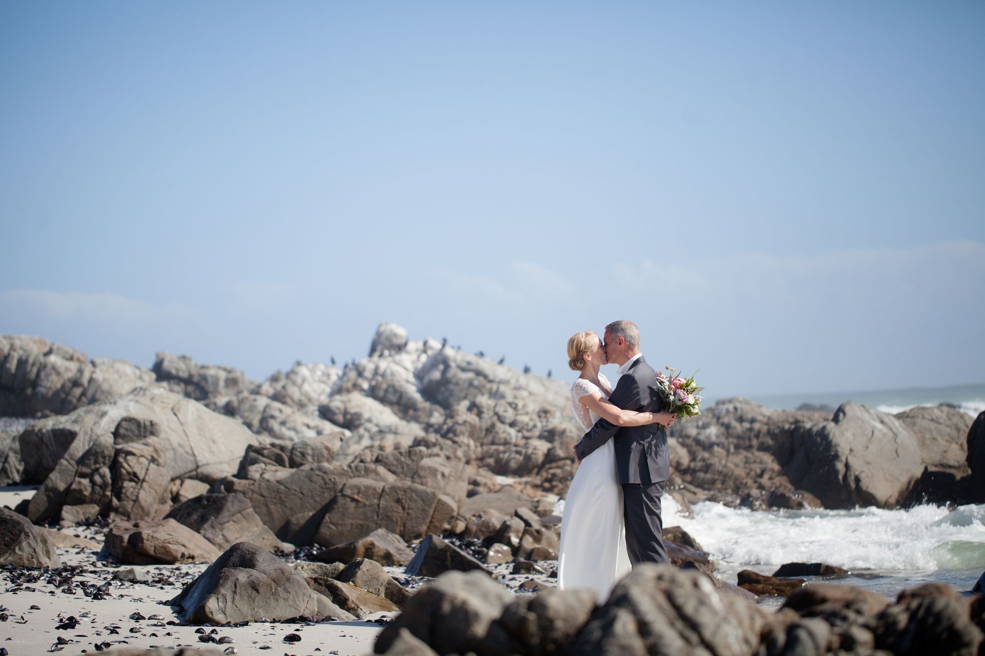A bride and groom are kissing on a rocky beach.