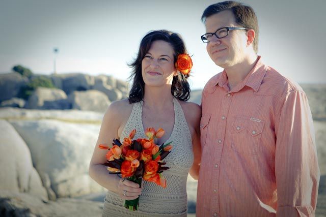 A man and a woman are posing for a picture on the beach . the woman is holding a bouquet of orange flowers.