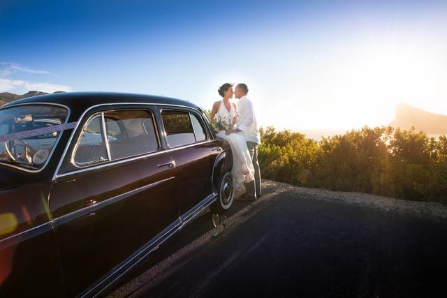 A bride and groom are kissing on the side of a car.