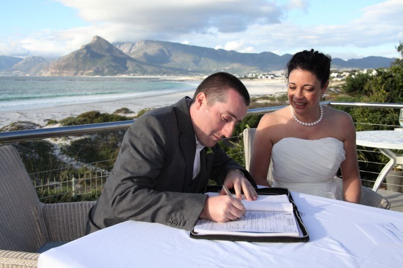 A bride and groom are signing a document on a balcony overlooking the ocean