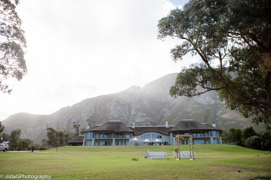 A large house sits on top of a grassy hill with mountains in the background
