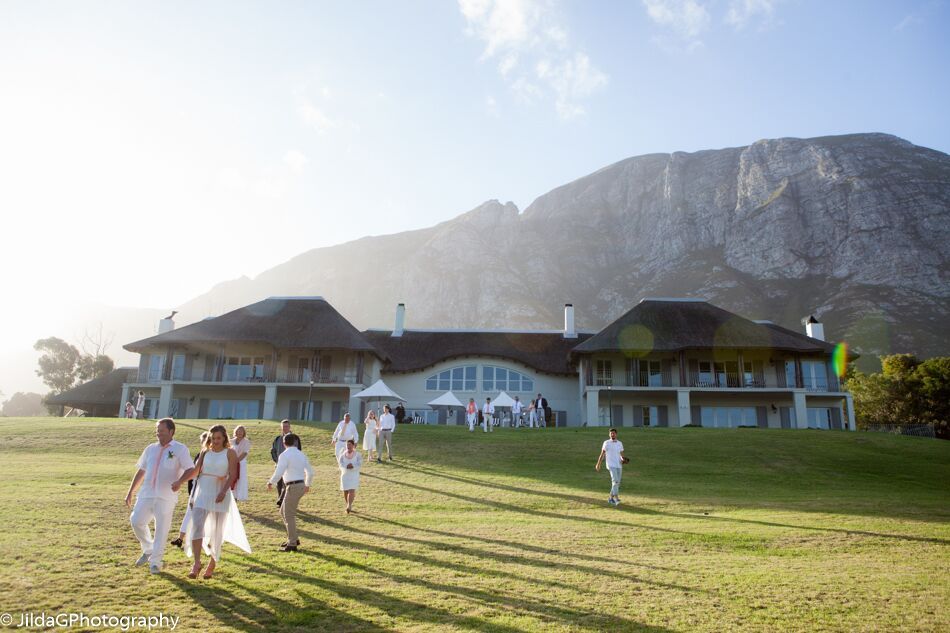 A group of people are walking in front of a large white house with mountains in the background.