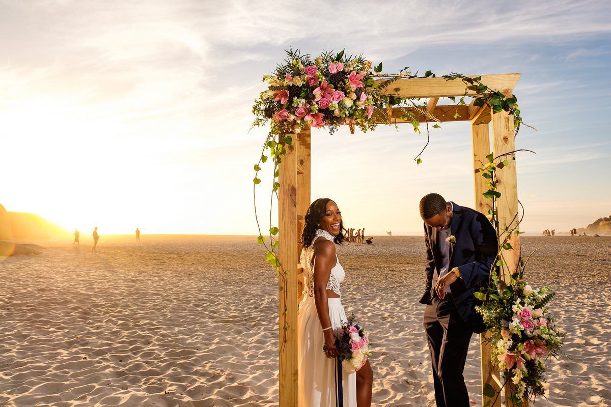 A bride and groom are standing under a wooden arch on the beach.