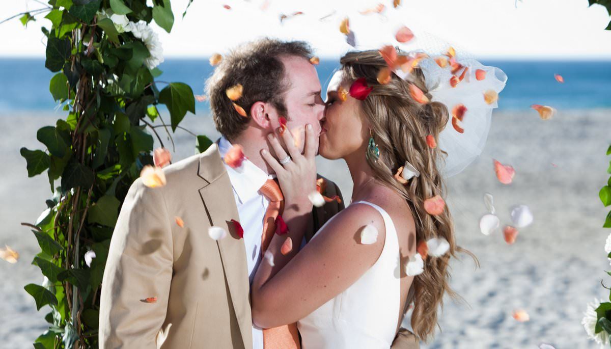 A bride and groom are kissing on the beach while confetti is being thrown at them.