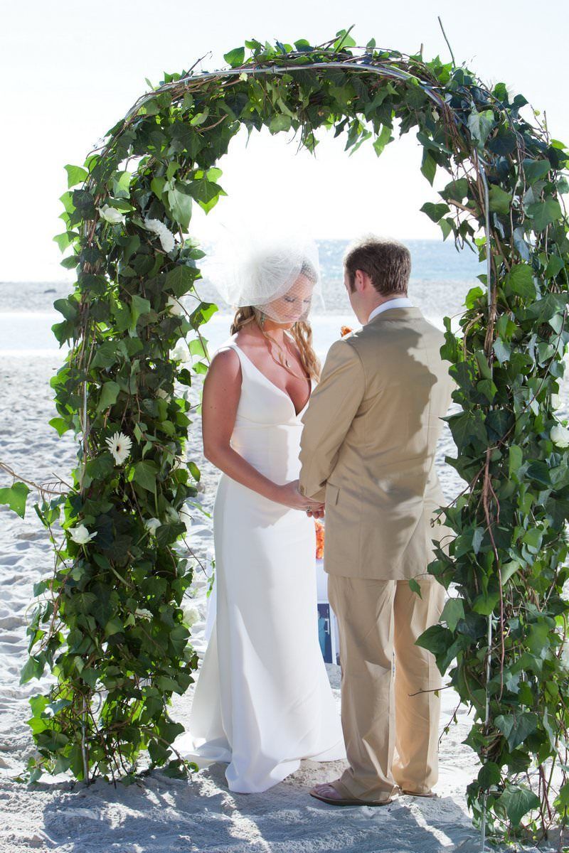 A bride and groom are holding hands under an arch on the beach.