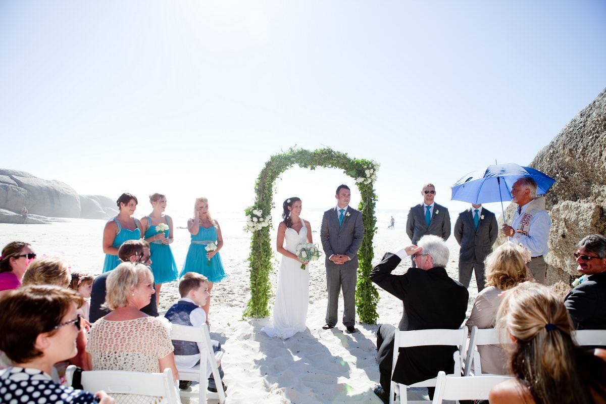 A bride and groom are getting married on the beach while their guests watch.