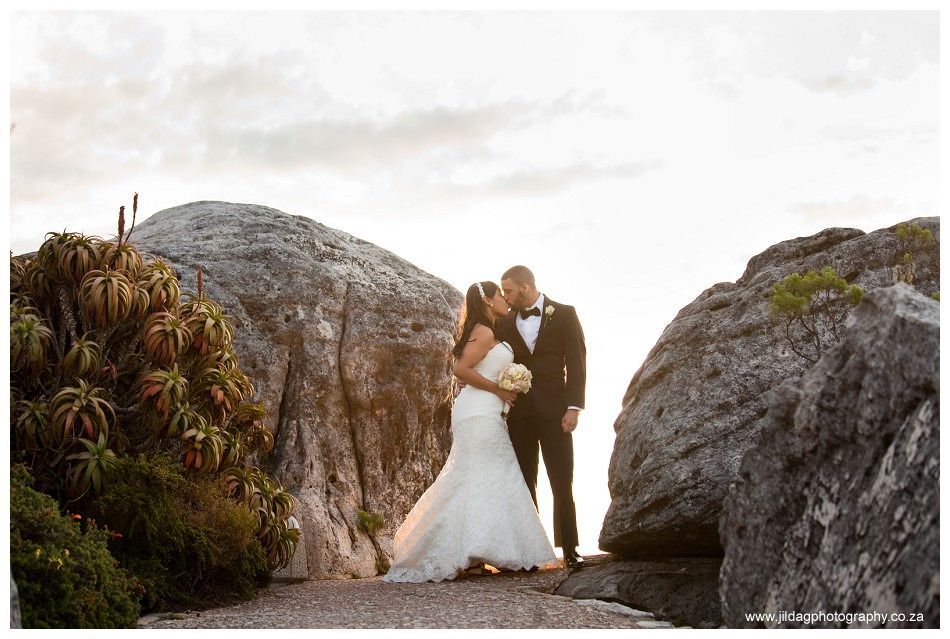 A bride and groom are standing next to each other on top of a rocky hill.